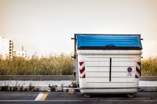 A large white industrial-sized skip with a blue hinged lid is positioned on a paved surface beside a curb in an outdoor urban setting near Canary Wharf. The skip has red and white chevron reflective markings on its sides and a small circular no-parking sign affixed to its front. Surrounding the skip, there is a strip of grass and weeds growing along the edge of the pavement, with a background comprising a clear sky and distant tall buildings typical of Canary Wharf's modern skyline. The scene suggests routine rubbish collection or waste removal activity, consistent with services offered by Commercial Waste Removal Canary Wharf, to manage commercial or construction debris efficiently in the local area. The lighting appears natural, indicating daytime, with the image capturing the functional and utilitarian environment common to rubbish removal operations in London’s financial district.