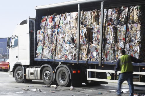 A large white commercial waste removal truck with an open cargo area filled with tightly packed compressed bales of mixed recyclable and non-recyclable waste materials, predominantly in shades of white, grey, and various colors from paper, plastic, and cardboard. The truck is parked on a paved industrial or commercial site, with a grey metal barrier running along the length of the cargo area for safety and containment. In the foreground, a worker wearing a high-visibility yellow vest and dark trousers is seen walking past the truck, approaching the loading area. The background features a clear sky, indicating daytime, and a portion of a red vehicle is visible on the left side of the image. The scene emphasizes professional waste management services typical of commercial rubbish removal, in a setting that may be associated with urban or business district environments such as Canary Wharf, where efficient disposal and recycling processes are crucial. Commercial Waste Removal Canary Wharf often handles similar waste collection operations, ensuring the removal of bulk rubbish from business premises or commercial properties within the local postcode area.