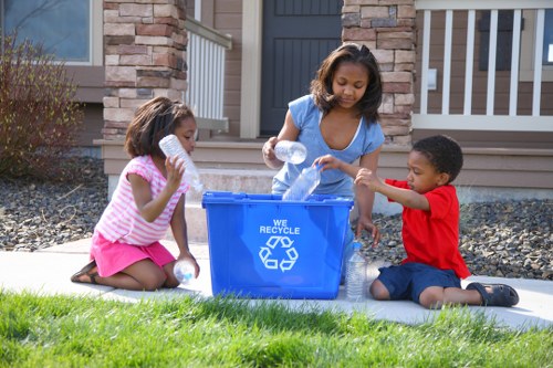 The image shows three children sitting on a concrete pathway in front of a residential building, engaging in recycling activities. The children, two girls and one boy, are placing empty plastic bottles into a blue recycling bin emblazoned with the recycling symbol and the words 'We Recycle.' The girl on the left, wearing a pink and white striped top and a pink skirt, has shoulder-length dark hair and is holding a clear bottle. The girl in the middle, with curly dark hair and wearing a light blue T-shirt, is actively placing a bottle into the bin. The boy on the right, dressed in a red T-shirt and dark shorts, is sitting cross-legged and reaching out to hand a bottle to the girl. The background features a house with stonework and a white porch railing, indicative of a suburban setting near Canary Wharf. The scene is well-lit with natural daylight, and the surrounding grass is green and well-maintained, emphasizing community engagement in waste sorting and recycling, consistent with rubbish removal services offered by Commercial Waste Removal Canary Wharf.