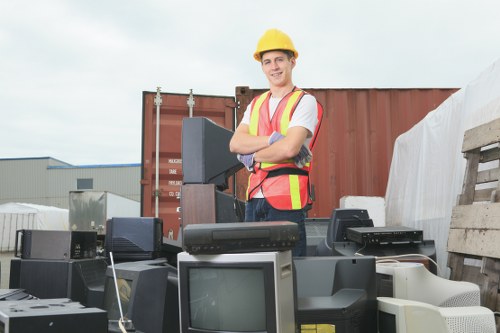 A young man wearing a yellow safety helmet and a high-visibility vest with orange reflective strips stands outdoors in an industrial or yard setting, behind a large collection of discarded electronic devices including old televisions, computer monitors, and desktop computers. The electronics are stacked and spread out on a surface, with some devices showing visible screens, buttons, and vents, mostly in black, grey, and white tones. In the background, there is a red shipping container, a wooden pallet, and some industrial buildings under an overcast sky. The environment appears to be a waste collection or rubbish removal site, consistent with professional rubbish disposal services provided by Commercial Waste Removal Canary Wharf, known for handling electronic waste or e-waste in the local Canary Wharf area near London. The scene suggests the process of clearing outdated electronics for proper disposal or recycling, relevant to the company’s rubbish removal operations in the area.