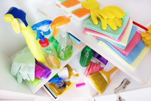 A collection of cleaning and rubbish removal supplies arranged on a white surface, including plastic spray bottles in blue, orange, and green, some with trigger nozzles, along with stacked colorful towels and sponges in pastel shades. Bright yellow cleaning gloves are placed on top of the towels. There are also small cleaning tools, such as a yellow-handled scrub brush and a small yellow dustpan. The items are organized in a way that suggests preparation for cleaning tasks, with some supplies leaning against each other and others placed neatly. The background is plain and white, emphasizing the vivid colors of the cleaning materials. This image reflects typical professional rubbish and cleaning supply arrangements, relevant to waste removal services offered in Canary Wharf or nearby areas, including the broader London postcodes. Commercial Waste Removal Canary Wharf handles such supplies during rubbish collection and disposal operations, with the scene situated on a clean and tidy workspace or storage area dedicated to cleaning equipment clearance.