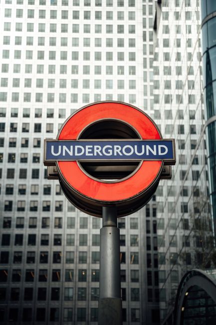 A London Underground station sign featuring a red circle and blue bar with the word 'UNDERGROUND' in white, mounted on a tall black pole in an urban setting. The sign is positioned in front of several