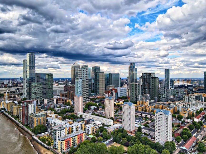 An expansive aerial view of a modern city skyline under a partly cloudy sky with a mix of tall glass skyscrapers and mid-rise office buildings. The foreground features a variety of residential apartme