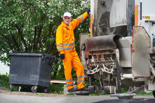 A waste collection worker dressed in bright orange high-visibility protective clothing with reflective stripes is standing on a paved pathway in an outdoor setting, near a large industrial rubbish compactor vehicle. The worker, who is wearing a white safety hat and black gloves, is positioned next to a black wheeled bin, which appears to be either full or waiting for collection. The rubbish compactor features metallic components, including a large roller and hydraulic mechanisms, and is situated adjacent to dense green foliage and trees, indicating a residential or commercial area in Canary Wharf. The scene is illuminated by natural daylight, highlighting the textures of the worker’s uniform, the smooth surface of the bin, and the industrial machinery’s metal finish. This image visually supports professional rubbish removal services offered by Commercial Waste Removal Canary Wharf, emphasizing efficient waste collection and environmental management in urban areas of London postcode areas such as E14 or nearby localities.