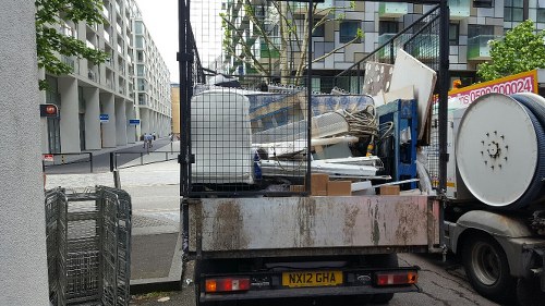 A flatbed truck parked on a city street in Canary Wharf, loaded with various discarded waste items including large white panels, metal frames, and plastic containers, arranged in a somewhat cluttered manner. The truck's metal surface shows signs of rust and wear, with some protective black mesh fencing enclosing part of the load. In the background, modern office buildings with glass facades and a street corner with traffic lights and pedestrian crossings are visible, indicating an urban environment. The scene suggests a waste removal operation by Commercial Waste Removal Canary Wharf, capturing typical rubbish collection activities for commercial properties within this business district, with minimal environmental impact on the surrounding area.